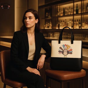 K&ouml;nnte beinhalten: Eine schwarze und cremefarbene Tragetasche mit einem bunten Truthahnmotiv. Die Tasche hat schwarze Griffe und eine schwarze Unterseite. Der Truthahn hat einen karierten K&ouml;rper und einen bunten Federf&auml;cher. Die Tasche steht auf einem braunen Hocker.