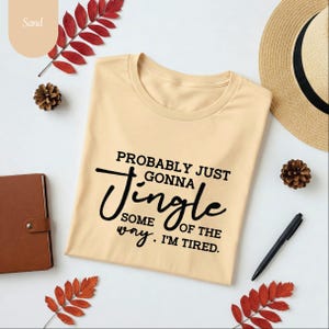 May include: Sand-colored t-shirt with the text "Probably Just Gonna Jingle Some of the Way. I'm Tired." The shirt is surrounded by a brown notebook, a pen, a straw hat, and decorative leaves and pinecones. The shirt is folded.