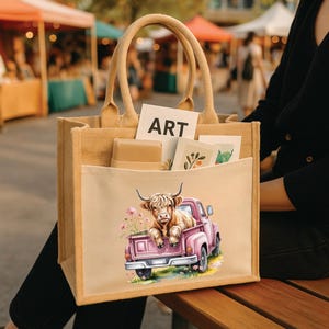 May include: A beige tote bag with a light pink pocket featuring a Highland cow in a pink pickup truck. The bag has a rectangular shape with two handles and the word "ART" is visible.