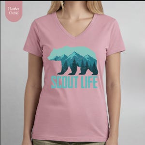 Puede incluir: Una camiseta en color rosa empolvado con cuello en V, con una silueta de oso turquesa con un dise&ntilde;o de monta&ntilde;as y bosques. El texto "SCOUT LIFE" est&aacute; debajo en turquesa. La camiseta tiene mangas cortas y un corte holgado.