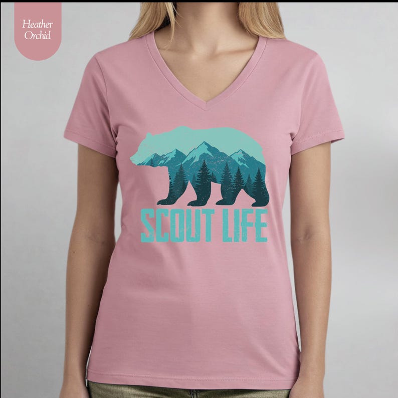 Puede incluir: Una camiseta en color rosa empolvado con cuello en V, con una silueta de oso turquesa con un dise&ntilde;o de monta&ntilde;as y bosques. El texto "SCOUT LIFE" est&aacute; debajo en turquesa. La camiseta tiene mangas cortas y un corte holgado.