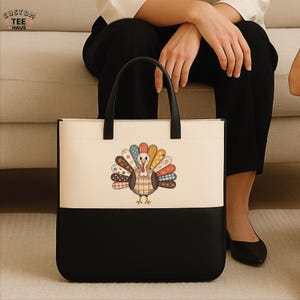 K&ouml;nnte beinhalten: Eine zweifarbige Tragetasche mit einem cremefarbenen oberen Bereich und einem schwarzen unteren Bereich. Die Tasche zeigt ein Truthahn-Design mit bunten Patchwork-Federn. Die Tasche hat schwarze Griffe und der Text "CUSTOM TEE HAUS" ist in der oberen linken Ecke sichtbar.