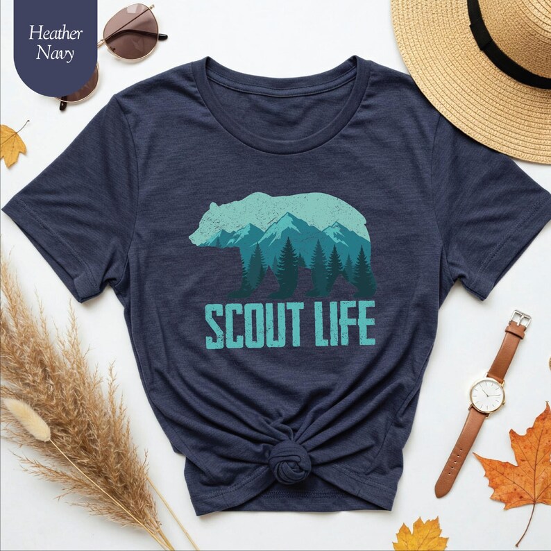Puede incluir: Camiseta azul marino jaspeado con un oso y un gr&aacute;fico de monta&ntilde;a. La silueta del oso est&aacute; rellena con una escena de monta&ntilde;a y bosque en tonos turquesa. Las palabras "SCOUT LIFE" est&aacute;n impresas debajo en turquesa.