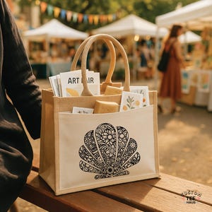 May include: A beige tote bag with a large black and white seashell design on the front. The bag has a light beige pocket and handles. The bag is filled with various items, including art prints and small packages. The text "ART IR" is visible.
