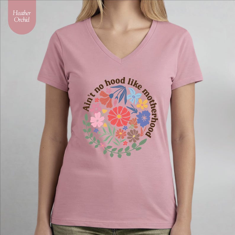 Peut inclure: T-shirt rose Heather Orchid &agrave; col en V avec l'inscription "Ain't no hood like motherhood" dans un motif floral circulaire. Le motif comprend diverses fleurs color&eacute;es et des feuilles vertes. Le t-shirt est en tissu doux.