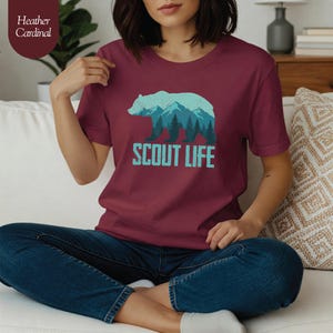 Puede incluir: Camiseta color cardenal con un dise&ntilde;o de oso y monta&ntilde;as en tonos azules y verdes, con el texto "SCOUT LIFE" debajo. La modelo lleva vaqueros azules. La camiseta es de color cardenal.