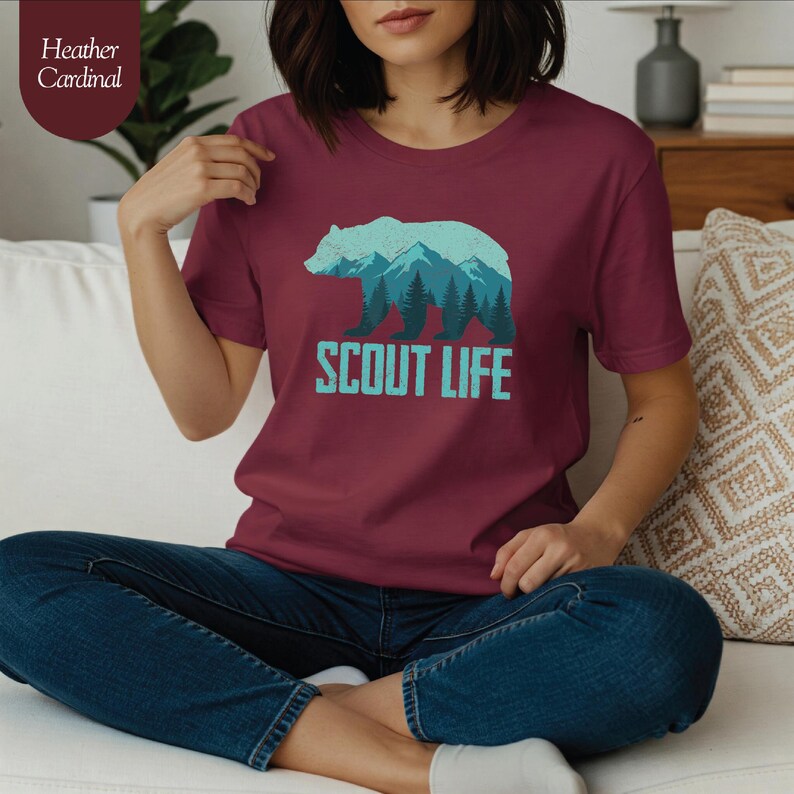 Puede incluir: Camiseta color cardenal con un dise&ntilde;o de oso y monta&ntilde;as en tonos azules y verdes, con el texto "SCOUT LIFE" debajo. La modelo lleva vaqueros azules. La camiseta es de color cardenal.