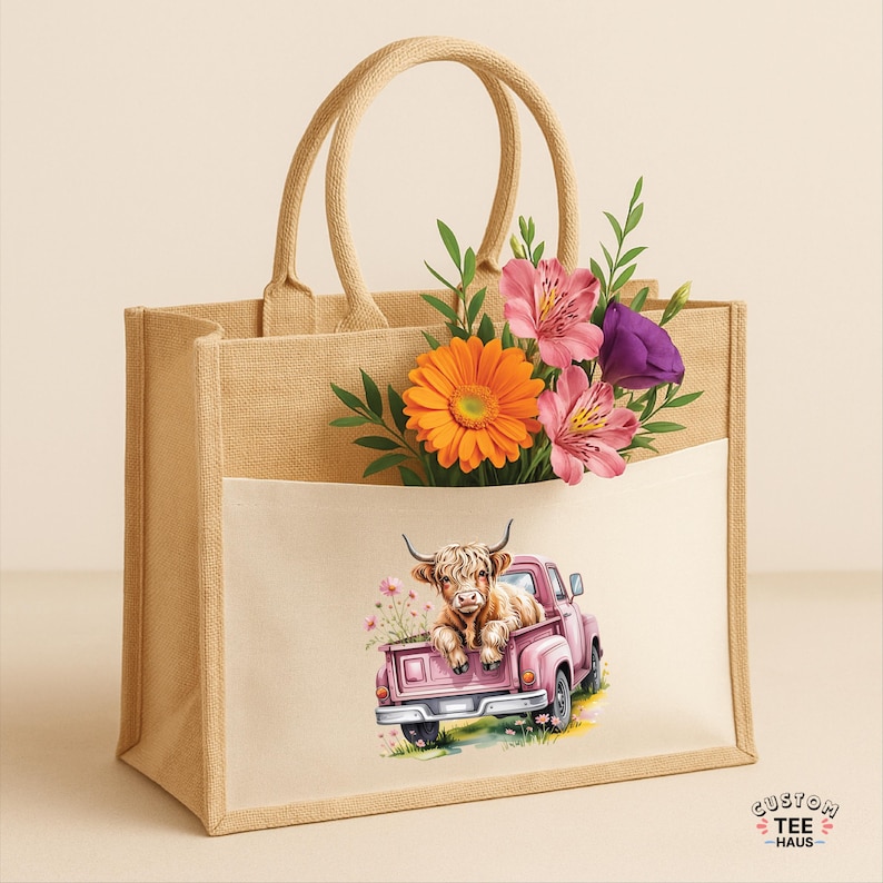 May include: A beige tote bag with a pocket featuring a watercolor illustration of a Highland cow in a pink pickup truck. The bag is filled with colorful flowers, including orange gerbera daisies and pink alstroemeria.