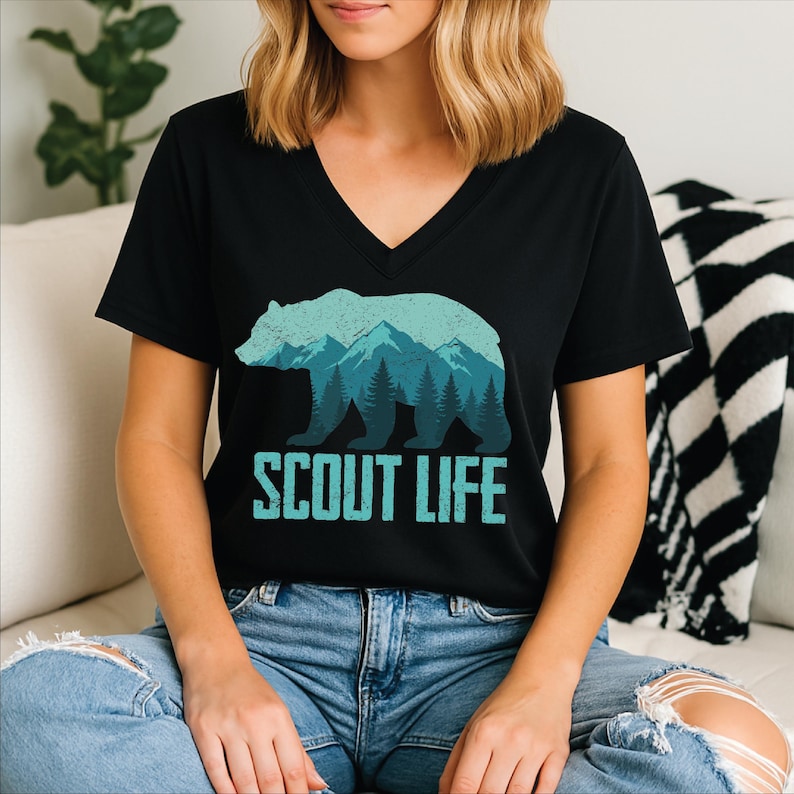 Puede incluir: Camiseta negra con cuello en V con un dise&ntilde;o de oso y monta&ntilde;a en tonos azules y turquesas. El texto "SCOUT LIFE" est&aacute; impreso debajo en turquesa. La camiseta es de material suave.