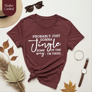 May include: A maroon t-shirt with the text "PROBABLY JUST GONNA JINGLE SOME OF THE WAY. I'M TIRED." in white script. The shirt is styled with sunglasses, a watch, and a bracelet. A straw hat and dried leaves are also in the image.