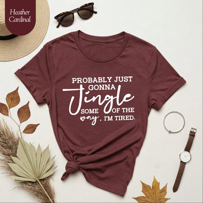 May include: A maroon t-shirt with the text "PROBABLY JUST GONNA JINGLE SOME OF THE WAY. I'M TIRED." in white script. The shirt is styled with sunglasses, a watch, and a bracelet. A straw hat and dried leaves are also in the image.