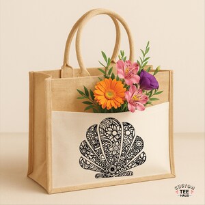 May include: A beige tote bag with a white pocket featuring a black seashell design filled with flowers. The bag has two handles and the text "CUSTOM TEE HAUS" is visible.