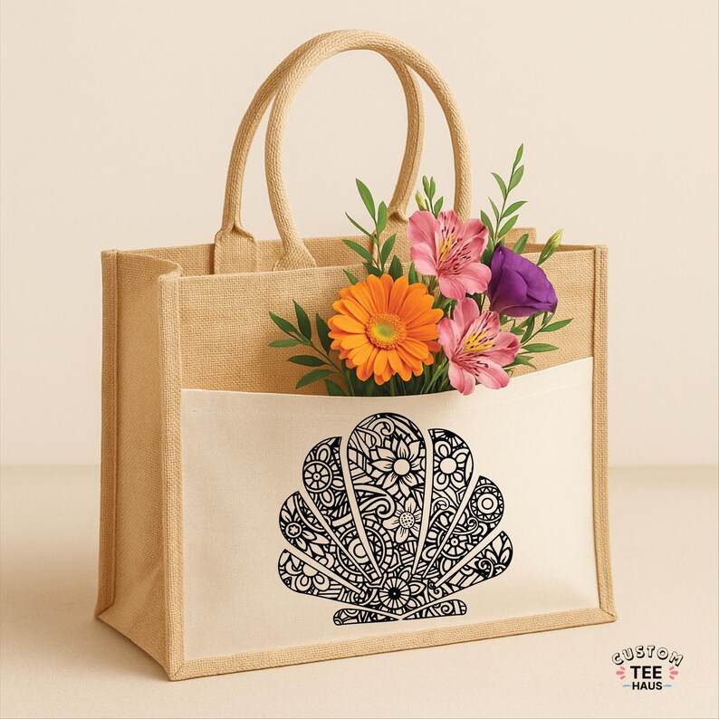 May include: A beige tote bag with a white pocket featuring a black seashell design filled with flowers. The bag has two handles and the text "CUSTOM TEE HAUS" is visible.