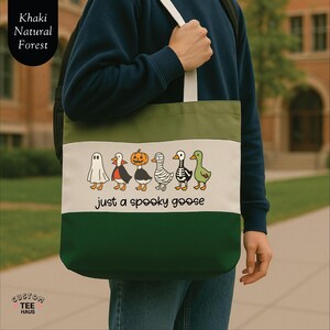Op de afbeelding: Een groene, witte en kaki tote bag met een Halloween-thema. De tas heeft een rij cartoon ganzen verkleed als een spook, vampier, pompoen, mummie, skelet en groene gans. De tekst "just a spooky goose" staat eronder.