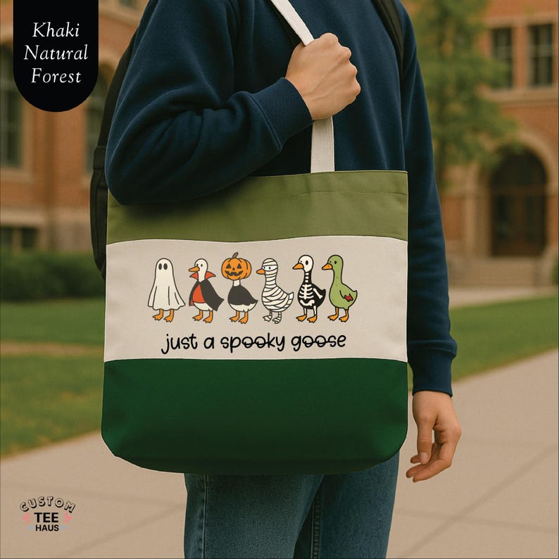 Op de afbeelding: Een groene, witte en kaki tote bag met een Halloween-thema. De tas heeft een rij cartoon ganzen verkleed als een spook, vampier, pompoen, mummie, skelet en groene gans. De tekst "just a spooky goose" staat eronder.