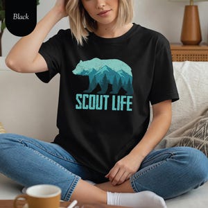 Puede incluir: Camiseta negra con un dise&ntilde;o de oso turquesa que incorpora monta&ntilde;as y &aacute;rboles. El texto "SCOUT LIFE" est&aacute; impreso debajo. La camiseta est&aacute; hecha de un material suave.