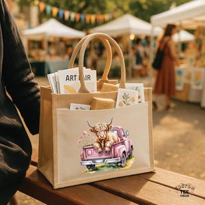 May include: A beige tote bag with a pink truck graphic carrying a Highland cow. The bag has a pocket and woven handles. The bag is filled with art prints and small packages. The words "ART IR" are visible on a print.