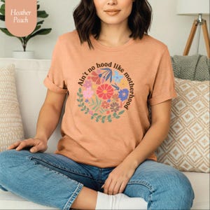 Peut inclure: T-shirt p&ecirc;che avec le texte "Ain't no hood like motherhood" dans un motif floral. Le t-shirt &agrave; manches courtes est fait d'une mati&egrave;re douce. Le t-shirt est de couleur p&ecirc;che.