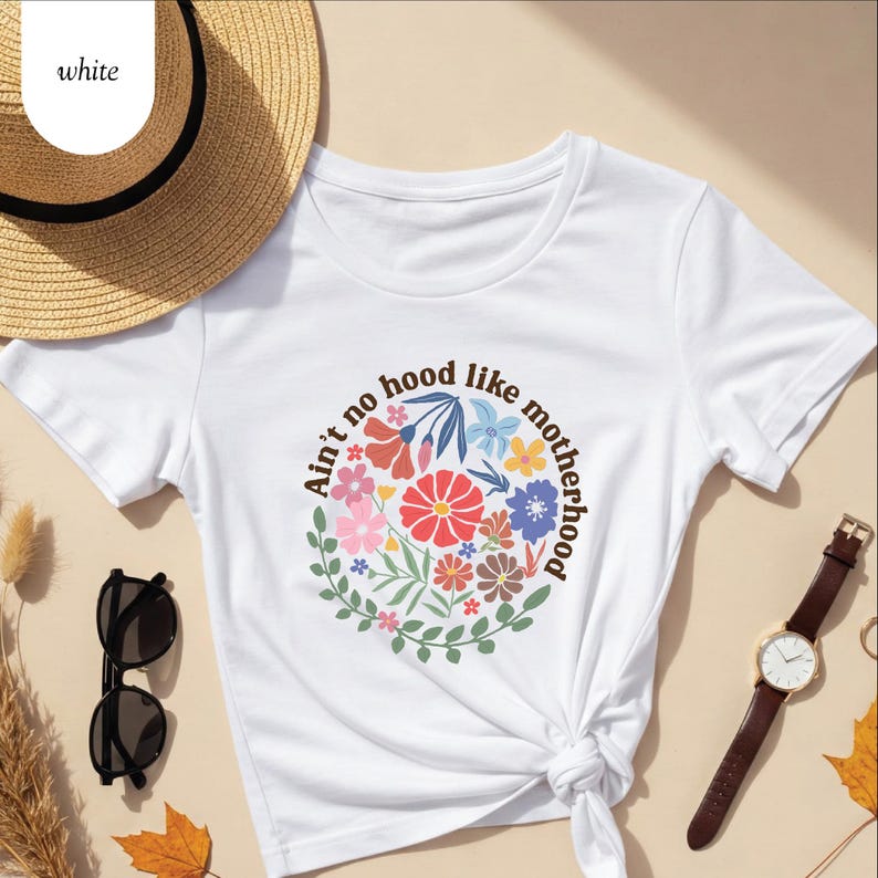 Peut inclure: T-shirt blanc avec un motif floral et le texte "Ain't no hood like motherhood" dispos&eacute; en cercle. Le motif pr&eacute;sente des fleurs color&eacute;es et des feuilles vertes. Le t-shirt est nou&eacute; &agrave; la taille.