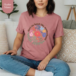 Peut inclure: T-shirt mauve chin&eacute; avec un motif floral et le texte "Ain't no hood like motherhood" dispos&eacute; en cercle. Le motif pr&eacute;sente des fleurs color&eacute;es et des feuilles vertes. Le t-shirt est &agrave; col rond et &agrave; manches courtes.