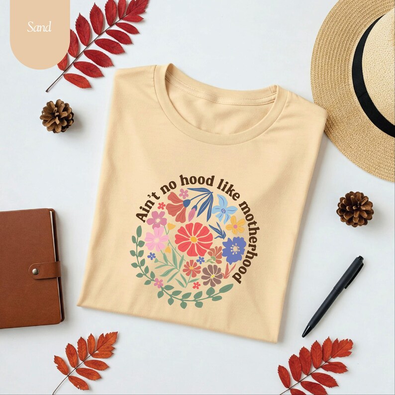 Peut inclure: Un t-shirt beige avec un motif floral et le texte "Ain't no hood like motherhood" dispos&eacute; en cercle. Le motif comprend diverses fleurs color&eacute;es et des feuilles vertes. Le t-shirt est pr&eacute;sent&eacute; avec un carnet, un stylo et des &eacute;l&eacute;ments d&eacute;coratifs.
