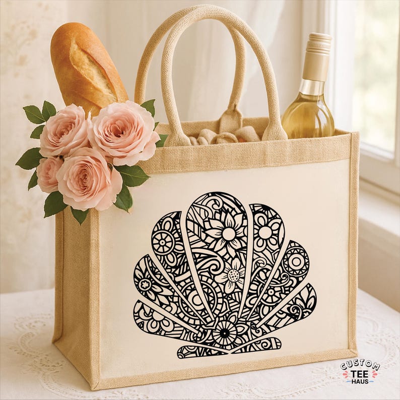 May include: A beige tote bag with a black seashell design, filled with a baguette, a bottle of wine, and pink roses. The bag has sturdy handles and is made of a natural-looking material. The bag is sitting on a white surface.