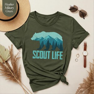Puede incluir: Camiseta verde oliva con un dise&ntilde;o de silueta de oso azul claro, con monta&ntilde;as y &aacute;rboles. El texto "SCOUT LIFE" est&aacute; impreso debajo del oso en azul claro. Un sombrero de paja y accesorios est&aacute;n en el fondo.