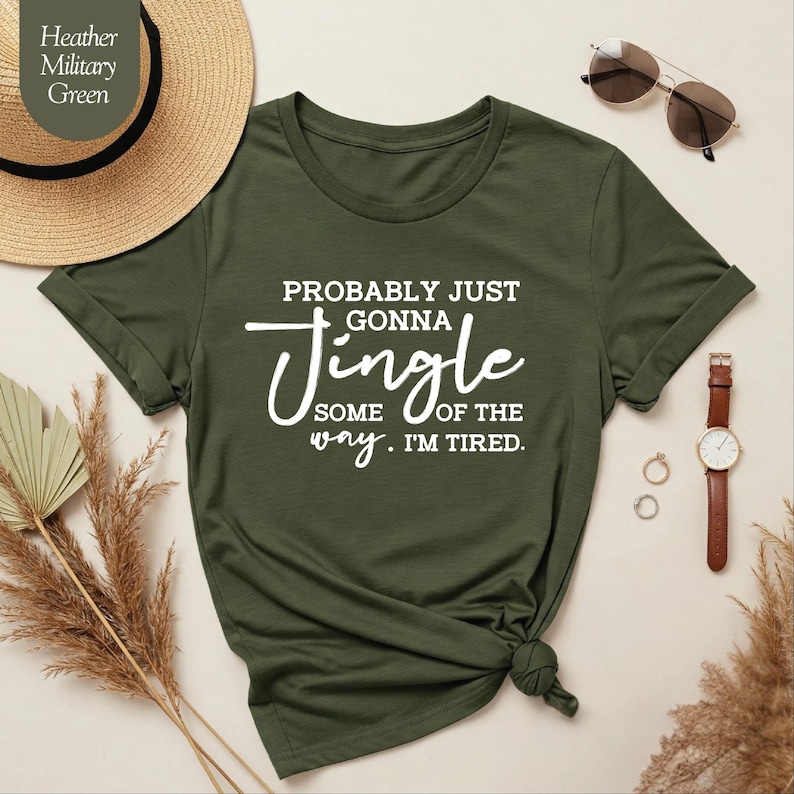 May include: Heather military green t-shirt with the text "Probably just gonna jingle some of the way. I'm tired." The shirt has rolled-up sleeves and a knotted hem. Accessories include a watch, sunglasses, and rings.
