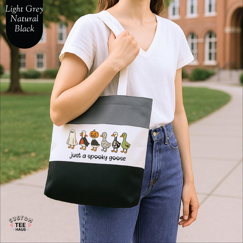 Op de afbeelding: Een canvas draagtas met een colorblock-ontwerp in lichtgrijs, naturel en zwart. De tas heeft een rij cartoon ganzen verkleed in Halloween kostuums met de tekst "just a spooky goose".