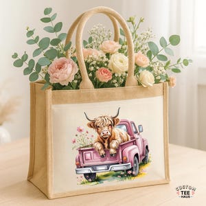 May include: A jute tote bag with a watercolor illustration of a Highland cow in a pink pickup truck. The bag has beige handles and is filled with pink and white flowers and greenery. The text "CUSTOM TEE HAUS" is in the lower right corner.