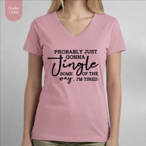 May include: A light pink V-neck t-shirt with the text "PROBABLY JUST GONNA Jingle SOME OF THE WAY. I'M TIRED." in black script. The shirt has short sleeves and is made of a soft material. The brand name "Heather Orchid" is in the upper left corner.