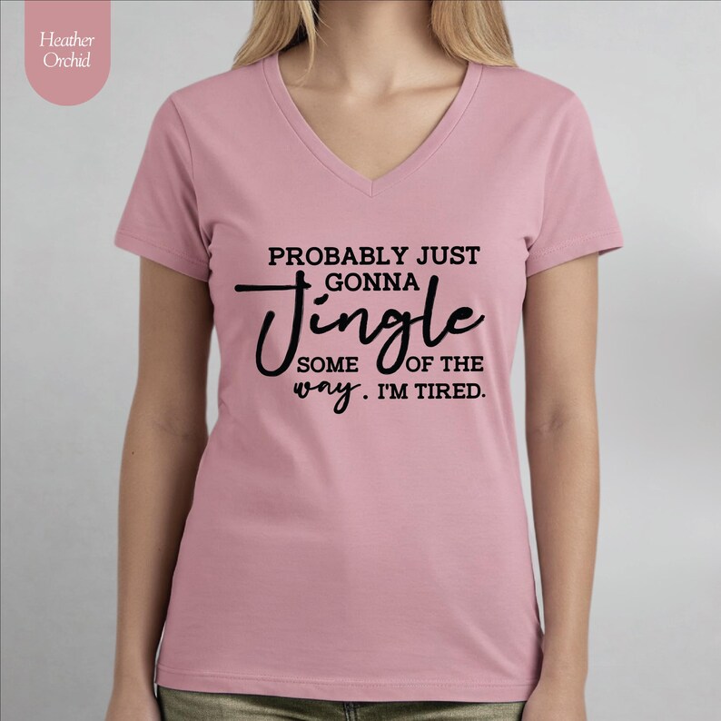 May include: A light pink V-neck t-shirt with the text "PROBABLY JUST GONNA Jingle SOME OF THE WAY. I'M TIRED." in black script. The shirt has short sleeves and is made of a soft material. The brand name "Heather Orchid" is in the upper left corner.