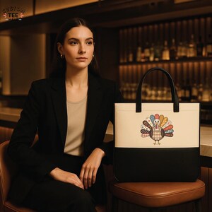 K&ouml;nnte beinhalten: Eine cremefarbene und schwarze Tragetasche mit einem Truthahnmotiv. Die Tasche hat schwarze Griffe und wird von einer Person mit einem schwarzen Blazer getragen. Das Truthahnmotiv weist eine Vielzahl farbenfroher Muster auf.