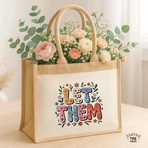May include: A beige jute tote bag with the words "LET THEM" in colorful, whimsical lettering. The bag is filled with a bouquet of pink and white roses, eucalyptus, and baby's breath. The bag has two beige handles.