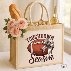 May include: A beige tote bag with the words "Touchdown Season" and a football graphic. The bag contains a baguette, a bottle of wine, and a bouquet of pink roses. The bag has two beige handles.