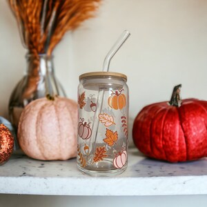 May include: A clear glass tumbler with a bamboo lid and a curved glass straw. The tumbler is decorated with fall-themed graphics, including pumpkins, leaves, and acorns in shades of orange, brown, and pink. The tumbler is sitting on a white surface.