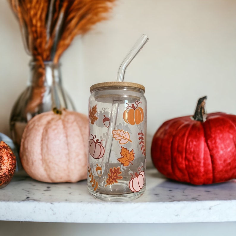 May include: A clear glass tumbler with a bamboo lid and a curved glass straw. The tumbler is decorated with fall-themed graphics, including pumpkins, leaves, and acorns in shades of orange, brown, and pink. The tumbler is sitting on a white surface.
