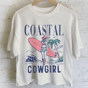 Op de afbeelding: Cr&egrave;mekleurig T-shirt met een afbeelding van een cowgirl met een surfboard, palmbomen en de woorden "Coastal Cowgirl Beach Club" in blauw. Het ontwerp heeft een vintage, verweerde uitstraling.