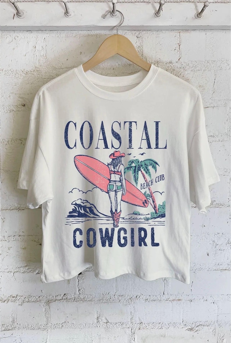 Op de afbeelding: Cr&egrave;mekleurig T-shirt met een afbeelding van een cowgirl met een surfboard, palmbomen en de woorden "Coastal Cowgirl Beach Club" in blauw. Het ontwerp heeft een vintage, verweerde uitstraling.