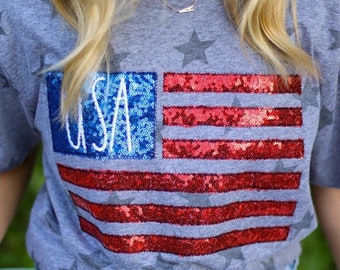May include: Gray t-shirt with a sequined American flag design. The flag features blue sequins with the word "USA" and red sequined stripes. The shirt also has gray star patterns.
