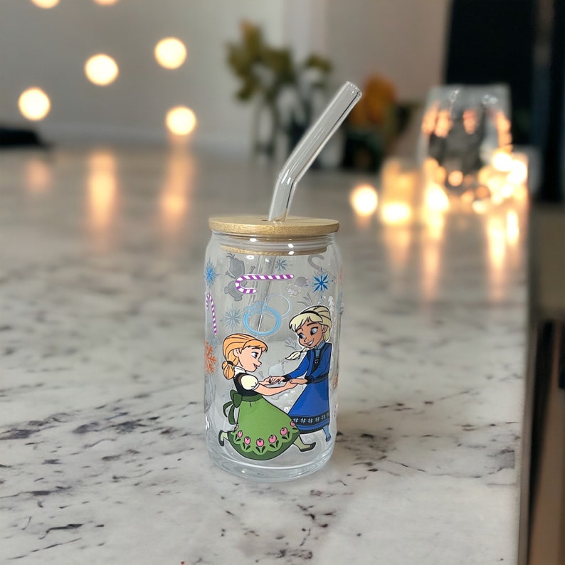 Puede incluir: Una lata de vidrio transparente con una tapa de madera y una pajita. La lata tiene un dise&ntilde;o de dos personajes de dibujos animados, una ni&ntilde;a con el pelo largo y rubio que lleva un vestido verde y un ni&ntilde;o con el pelo corto y rubio que lleva una camisa azul. Los personajes se dan la mano y hay copos de nieve y bastones de caramelo en el fondo.