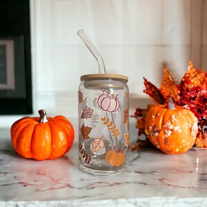 May include: A clear glass tumbler with a bamboo lid and a curved glass straw. The tumbler is decorated with fall-themed illustrations of pumpkins and leaves in shades of orange, brown, and pink. Two orange pumpkins are on either side of the tumbler.