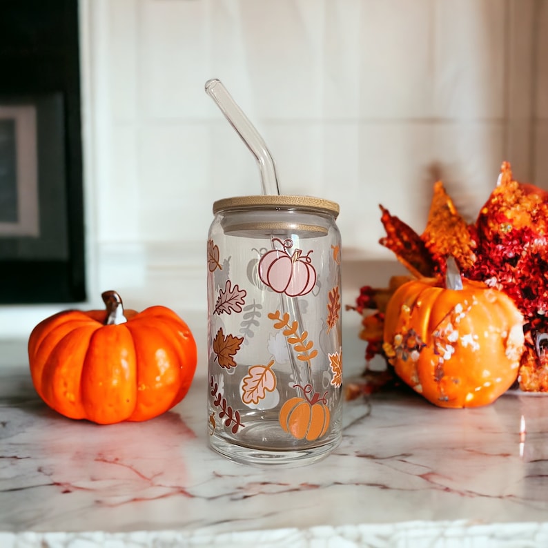 May include: A clear glass tumbler with a bamboo lid and a curved glass straw. The tumbler is decorated with fall-themed illustrations of pumpkins and leaves in shades of orange, brown, and pink. Two orange pumpkins are on either side of the tumbler.