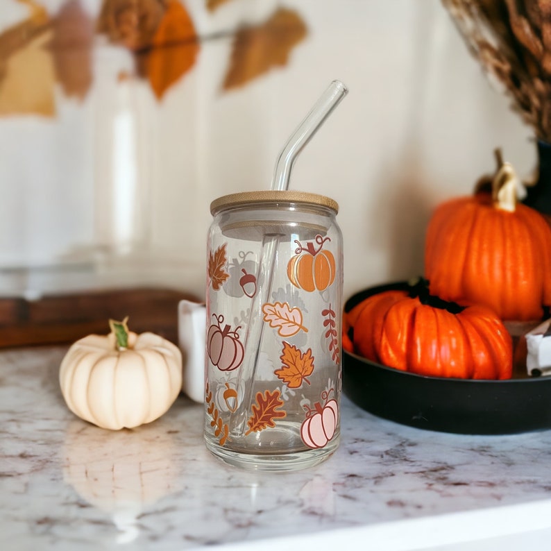 May include: A clear glass tumbler with a bamboo lid and a glass straw. The tumbler is decorated with orange, pink, and brown fall-themed graphics, including pumpkins and leaves. A white pumpkin and orange pumpkins are in the background.