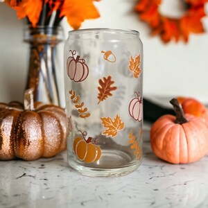 May include: A clear glass can-shaped tumbler decorated with fall-themed graphics. The design includes pumpkins, leaves, and an acorn in shades of orange, brown, and pink. The tumbler is displayed with pumpkins and fall foliage.