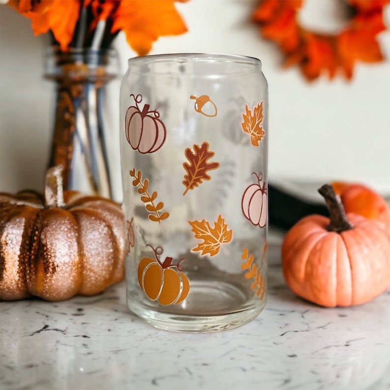 May include: A clear glass can-shaped tumbler decorated with fall-themed graphics. The design includes pumpkins, leaves, and an acorn in shades of orange, brown, and pink. The tumbler is displayed with pumpkins and fall foliage.