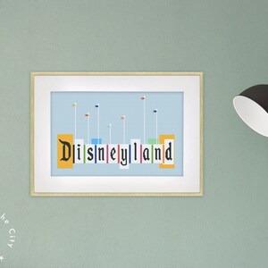 Puede incluir: Una impresi&oacute;n enmarcada de una ilustraci&oacute;n estilizada del letrero de Disneyland con banderas de colores. El letrero est&aacute; formado por bloques individuales con las letras "Disneyland" en negro.