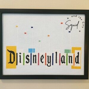 Puede incluir: Una obra de punto de cruz enmarcada que presenta la palabra "Disneyland" en letras negras con bloques de colores detr&aacute;s de cada letra. El texto "and the Squid City" est&aacute; en la esquina superior derecha.