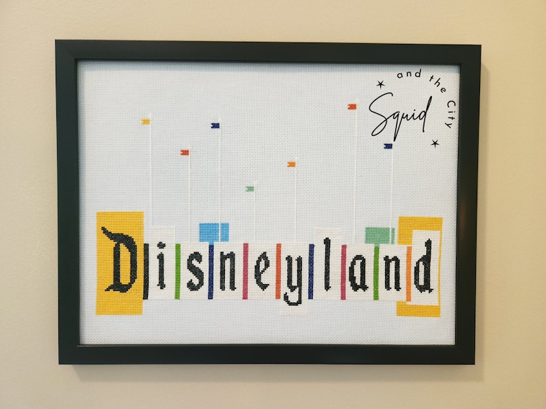 Puede incluir: Una obra de punto de cruz enmarcada que presenta la palabra "Disneyland" en letras negras con bloques de colores detr&aacute;s de cada letra. El texto "and the Squid City" est&aacute; en la esquina superior derecha.