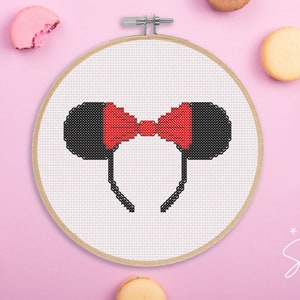 May include: A white cross-stitch embroidery hoop with a black and red Mickey Mouse ears headband design. The headband has a red bow in the center.